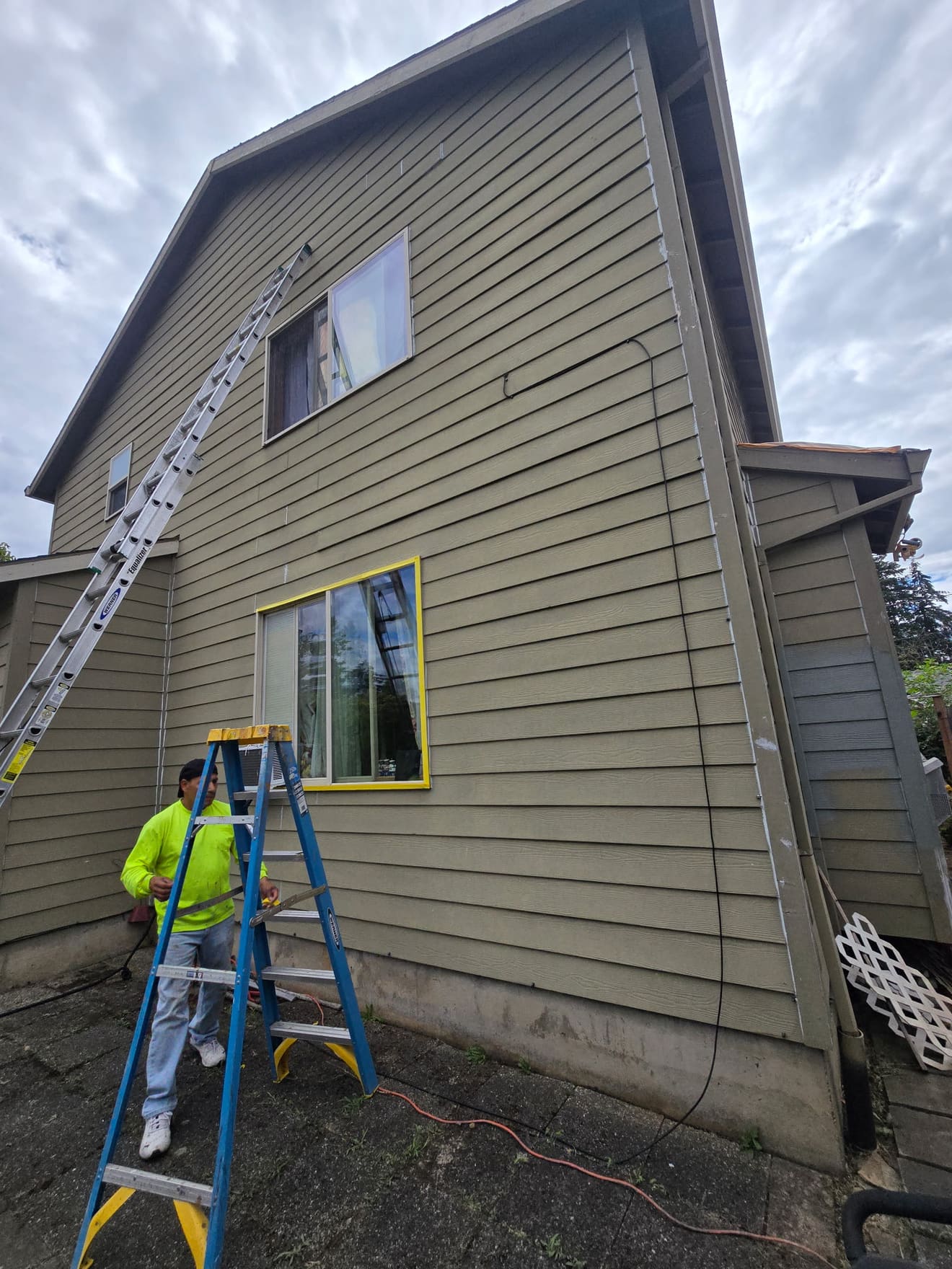 Two-story side wall before repaint — worn olive siding with ladder set up
